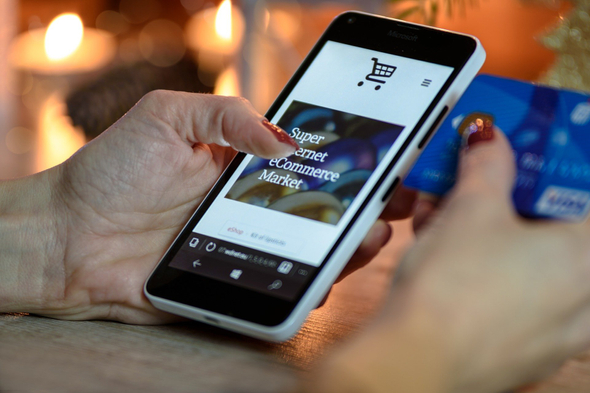 Na imagem é possível ver as mãos de uma mulher de meia idade com unhas vermelhas: a direita segurando um cartão azul, e na esquerda um aparelho smartphone acessando uma loja virtual.