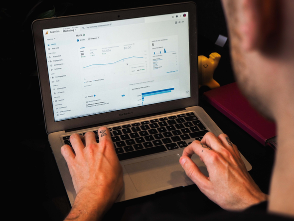 A imagem mostra um homem branco, usando o computador para acessar o google analytics, uma ferramenta importante na análise de dados. Em sua tela podemos observar o home da página, que apresenta alguns gráficos de acesso, valor e tempo.