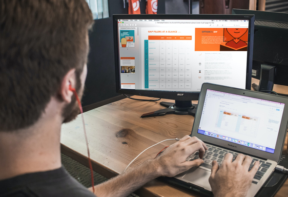 A imagem mostra um homem sentado em frente a um notebook cinza e um monitor preto. Na tela do monitor, é possível ver algumas ilustrações, que podem ser enviadas no e-mail marketing.