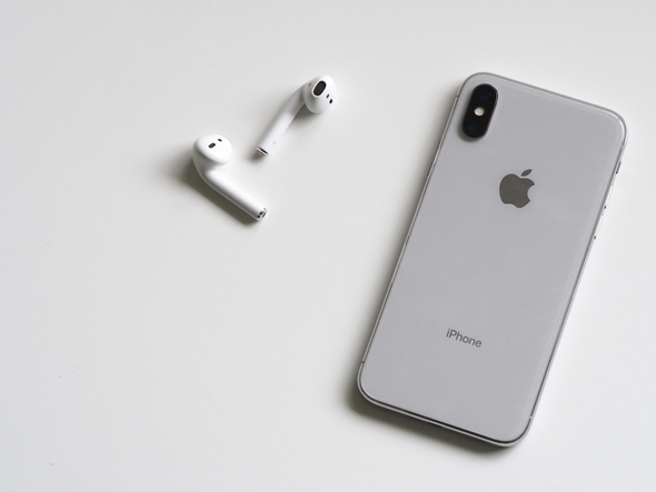A imagem mostra um aparelho smartphone iPhone prata e dois fones de ouvido bluetooth brancos deitados sobre uma superfície branca.