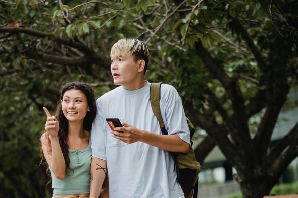 A imagem traz dois personagens, um homem e uma mulher asiáticos, numa área urbana com presença de árvores. A mulher, que tem uma blusa verde água, indica com os dedos algum lugar para o homem, que tem um smartphone em mãos.