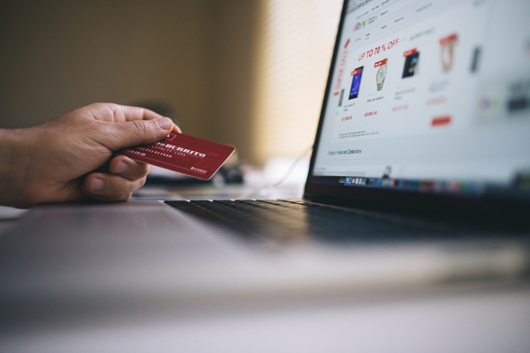 A imagem mostra um notebook aberto numa loja virtual com alguns produtos em promoção sendo exibidos. Também é possível ver uma mão segurando um cartão de crédito vermelho.