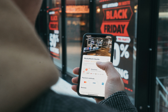 Pessoa segurando um smartphone em frente à uma loja que exibe promoções de Black Friday. Ela está concluindo a aquisição de algum produto ou serviço.