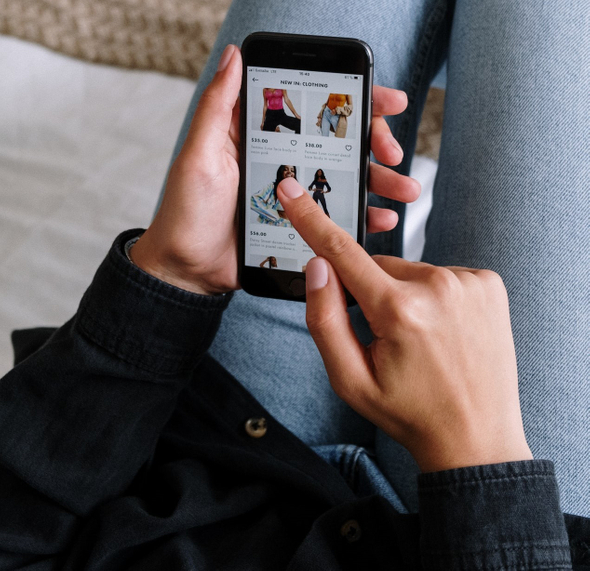 A imagem mostra uma pessoa acessando uma loja de roupas online através de um iPhone preto. Ela está segurando o celular com a mão esquerda e com o dedo indicador da mão direita navega pela página do e-commerce.