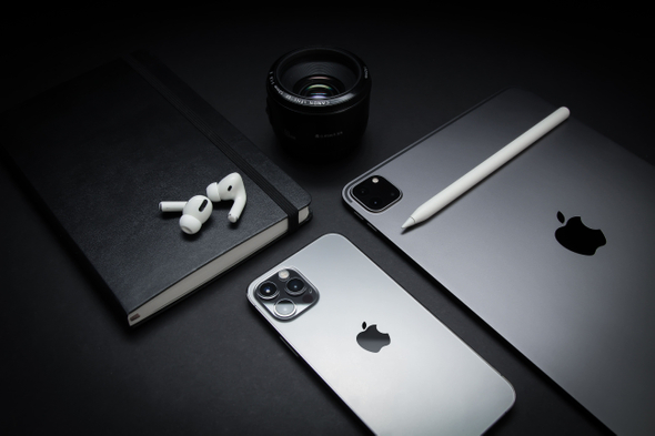 Sobre uma mesa preta, produtos da Apple como Iphone, Ipad, Airpods e uma lente de câmera HD, além de um bloco de anotações.
