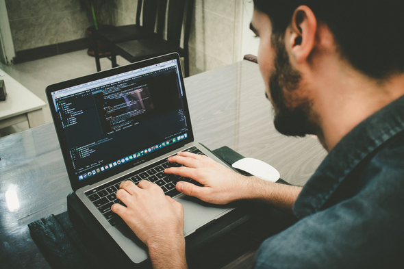 A imagem mostra um homem branco, com cabelos e barba pretos, sentado em frente a um notebook. Ele aparece de costas para a imagem, mexendo no computador. Na tela, é possível ver alguns códigos de programação.