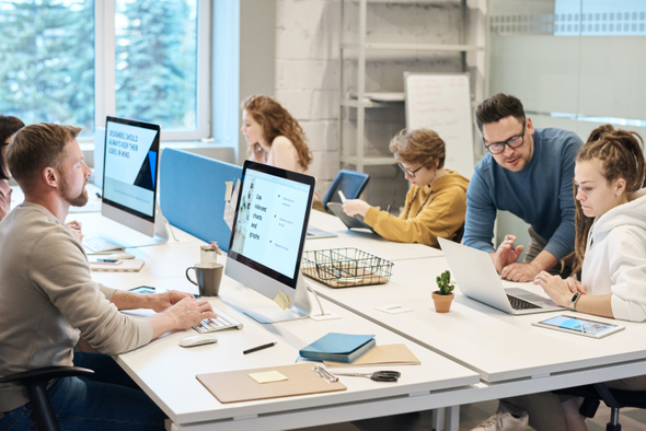 Na imagem observamos um escritório, onde algumas pessoas trabalham em computadores desktop, e no canto da imagem há um homem que parece estar ajudando uma mulher que trabalha em um notebook.