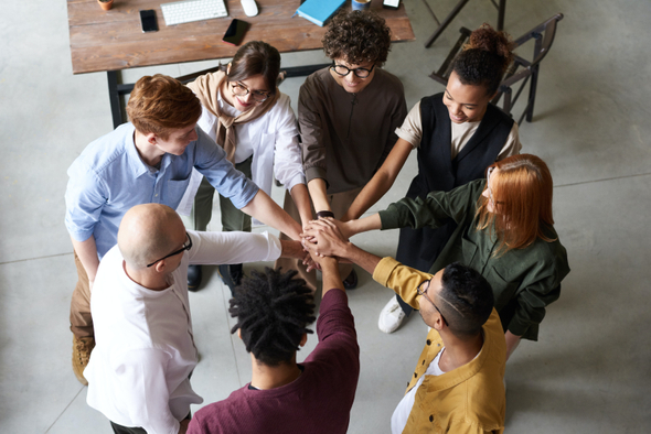 O ambiente da imagem é um escritório, nela há oito pessoas estão em roda, unindo as mãos no centro, como forma de comemorar o trabalho em equipe.