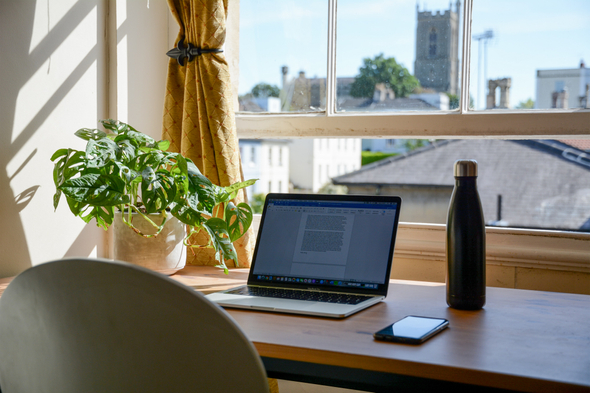 A imagem mostra uma mesa de madeira que se localiza em frente a uma janela, em cima dessa mesa há um notebook com um texto aberto e ao lado esquerdo dele há um vaso de planta e ao lado direito uma garrafa de água e um celular.