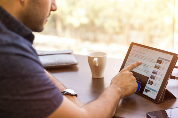 A imagem mostra um homem branco sentado em frente a uma mesa de escritório. Com a mão direita, ele mexe em um tablet, que está com a página aberta na área de edição de artigos do LinkedIn. 