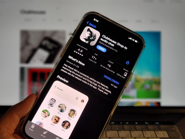 Alt text: A imagem mostra um aparelho iPhone na Apple Store, aberto na página de download do clubhouse. Ao fundo, é possível ver desfocada a tela de um computador e parte de seu teclado.