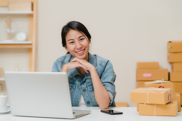 A imagem mostra uma mulher com traços asiáticos sentada em frente ao computador. Ela está olhando para a câmera e sorrindo. Ao lado dela, é possível ver diversas caixas de encomenda.