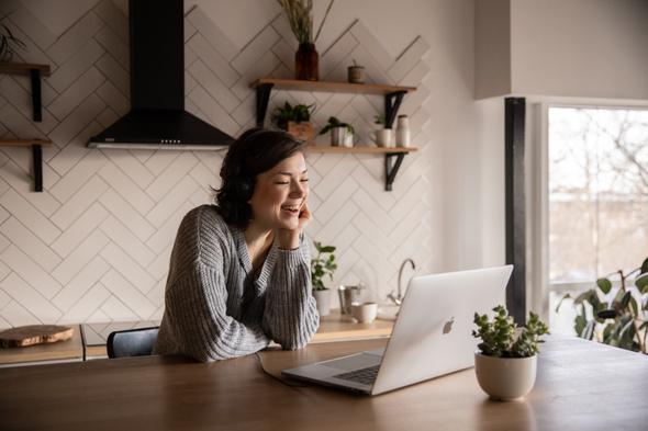 A imagem mostra uma mulher branca que está em home office, ela está sorrindo para o laptop prata que está a sua frente sobre uma mesa de madeira. Ao lado do computador há um vaso de planta.