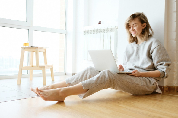 Mulher loira de cabelos curtos está sentada no chão mexendo no notebook. Ela veste uma blusa de moletom cinza e calças de pijama claras.