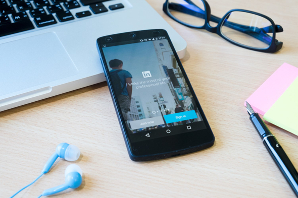 A imagem mostra um celular preto que está sob uma mesa de madeira. Na tela do aparelho, é possível ver a página inicial do app do LinkedIn, rede social que pode ser utilizada para o marketing. 